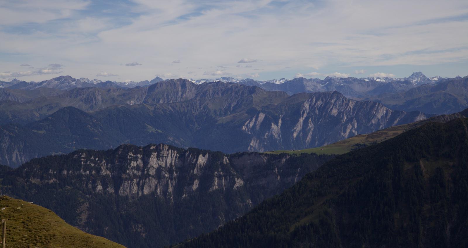 a view across a mountainous landscape; one pine forest slope in the foreground off to the right, one prominent light grey cliff in the centre, two or three layers of forest covered mountains, and finally black rock and white snow capped mountains along the horizon with a thin covering of white clouds