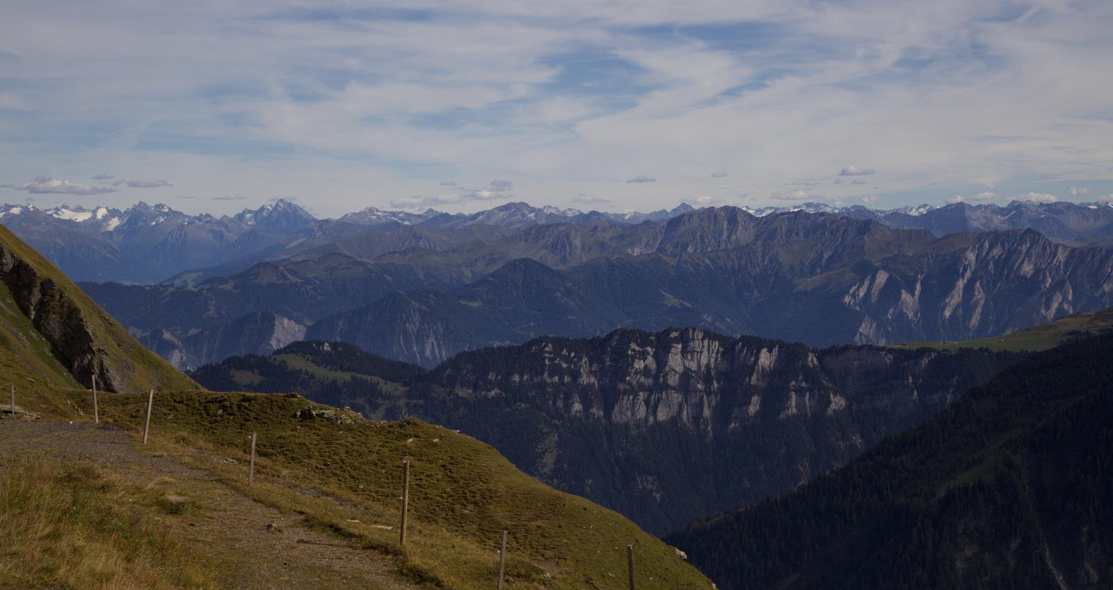 a view across a mountainous landscape; a grassy foreground with a small wire fence, then a few layers of rocky pine forest covered mountains, and finally black rock and white snow capped mountains along the horizon with a thin covering of white clouds
