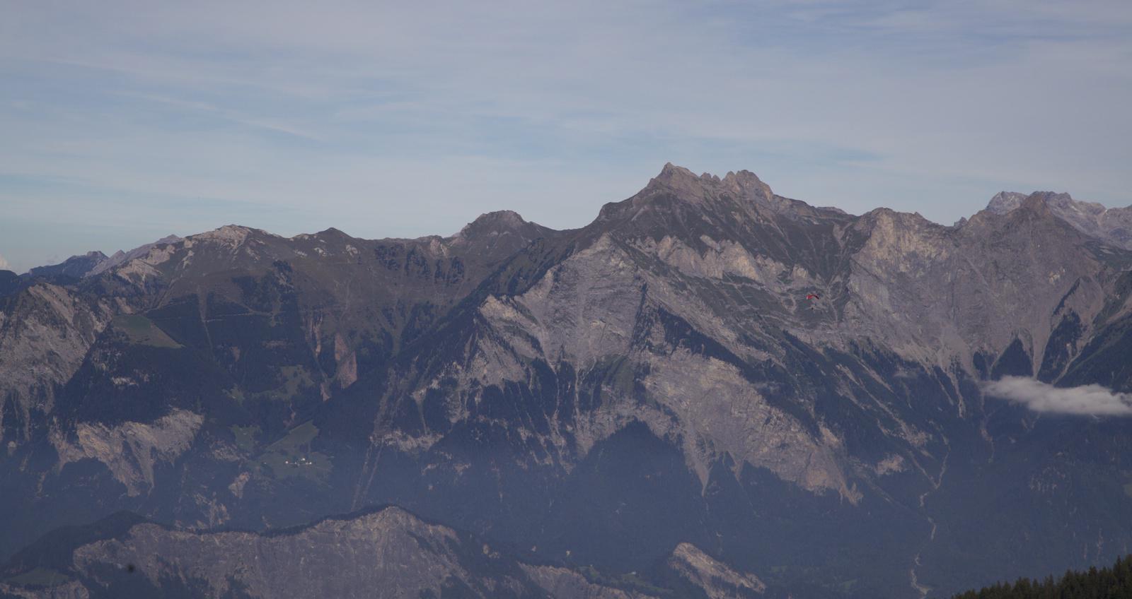 a far away hang glider shown against a massive mountainous backdrop with a greyish sky of whispy clouds